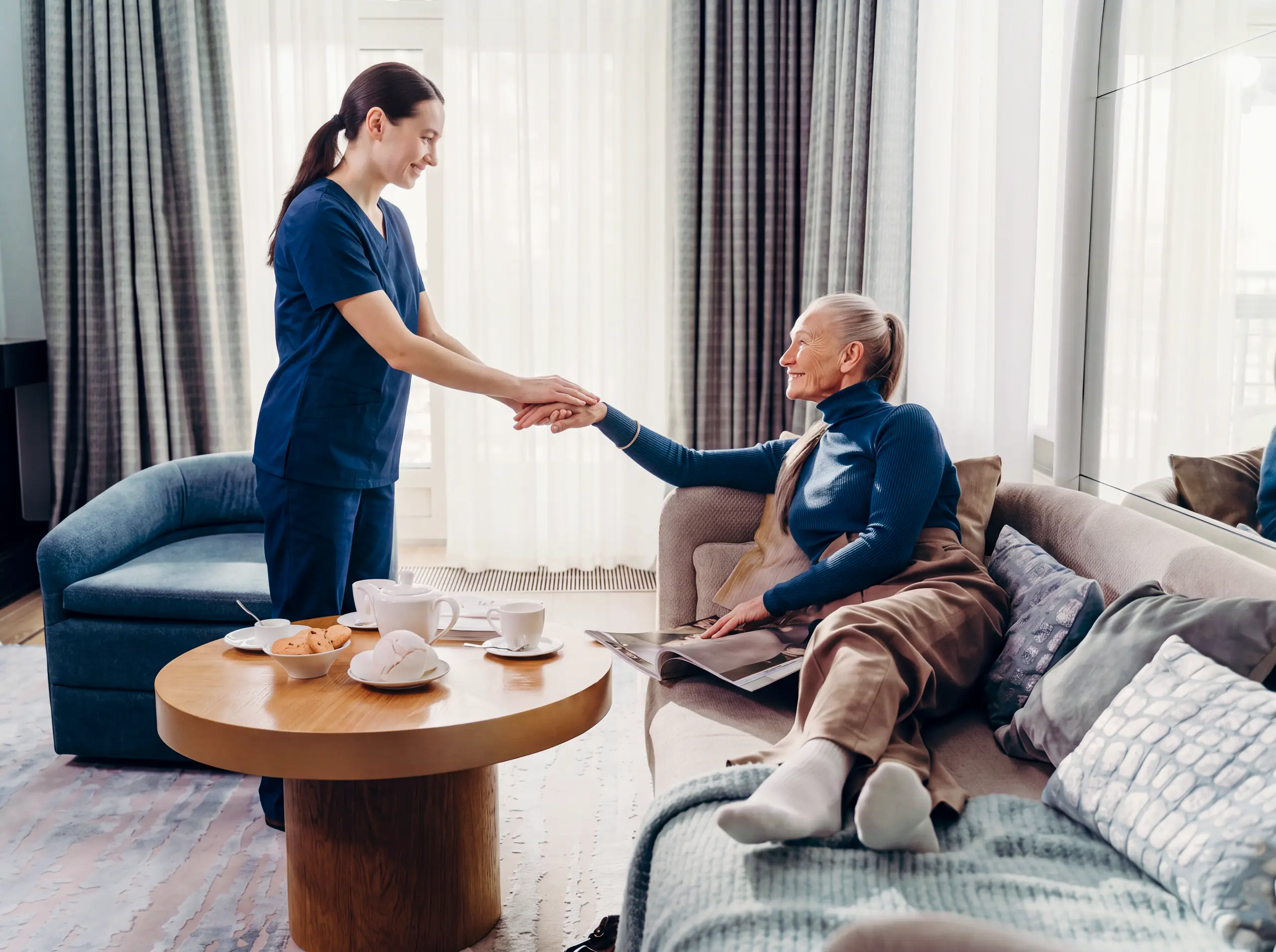 Caregiver in scrubs warmly holds hands with a smiling senior woman relaxing on her living room sofa.