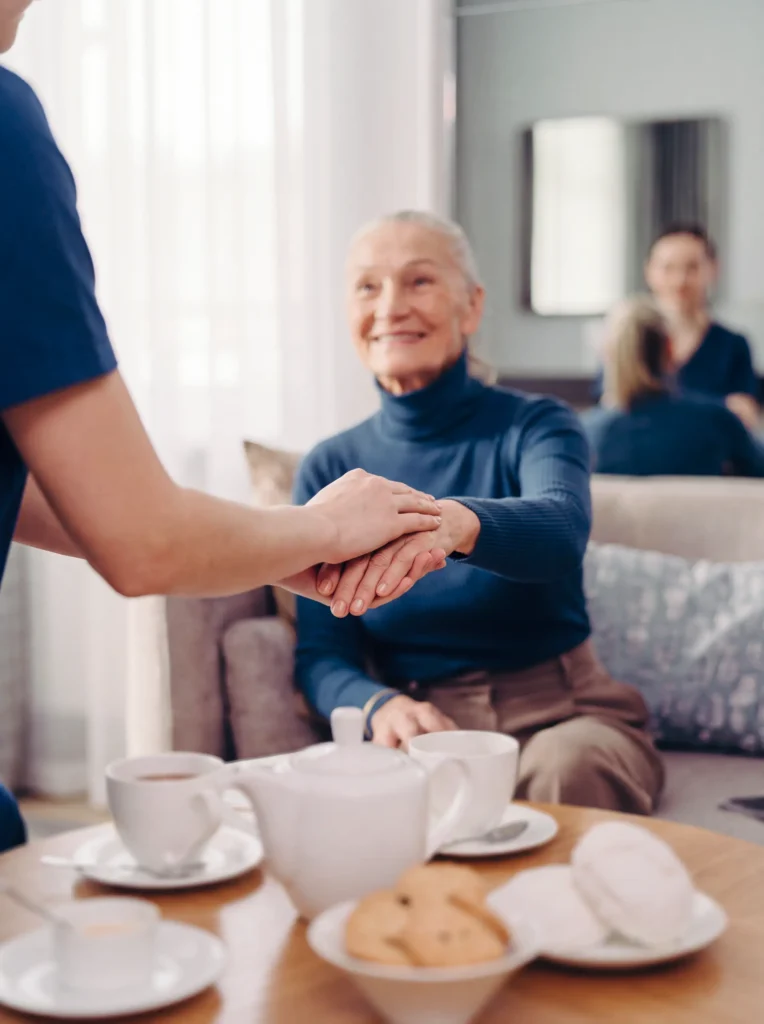 Caregiver gently holding a smiling senior woman's hands during tea time in her living room.
