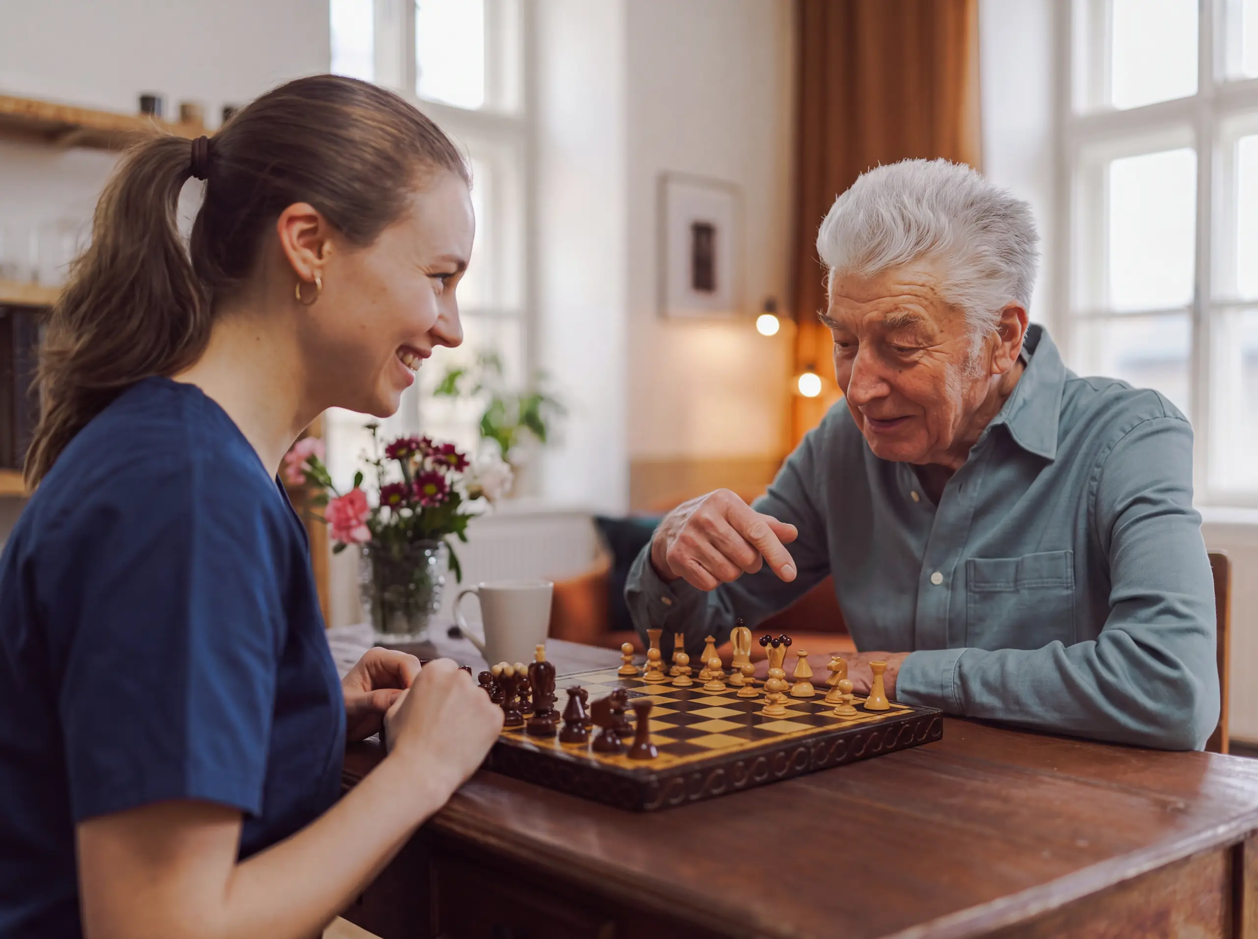 Smiling caregiver plays chess with a senior man at a wooden table in his bright living room.