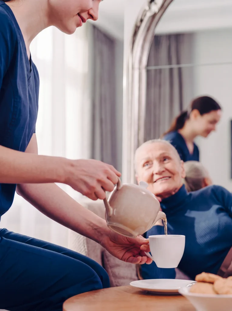 Caregiver in navy scrubs pours tea for a smiling senior woman in her living room.
