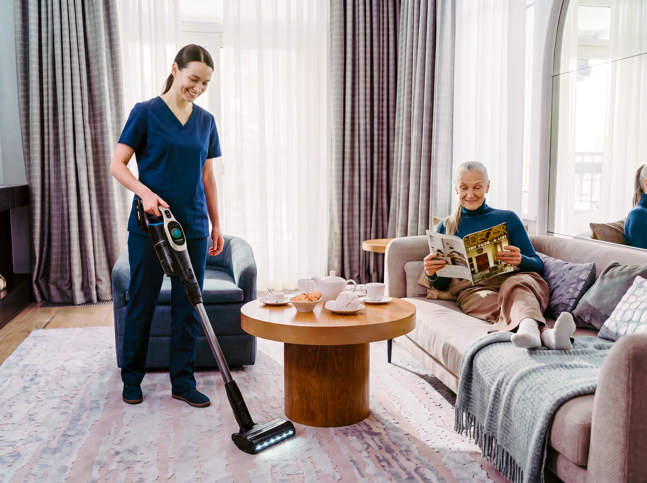 A smiling caregiver vacuums the living room rug while a senior woman relaxes on the sofa reading a magazine.