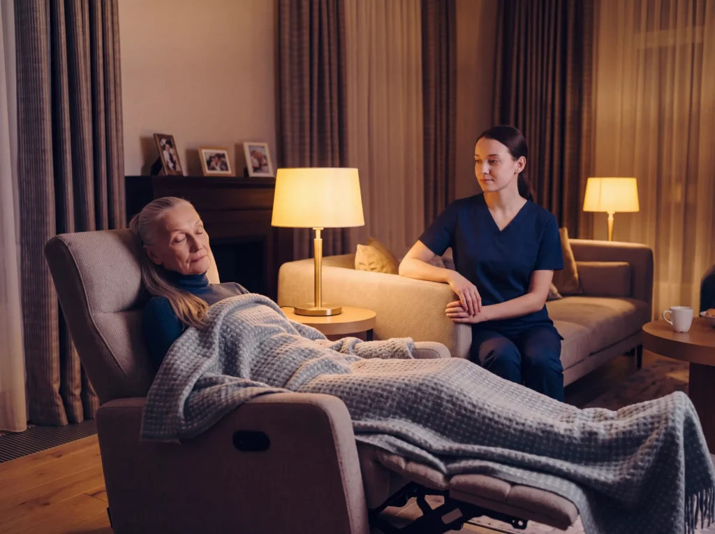 Caregiver sits beside a senior woman resting under a blanket in a recliner in a cozy, lamp-lit living room.