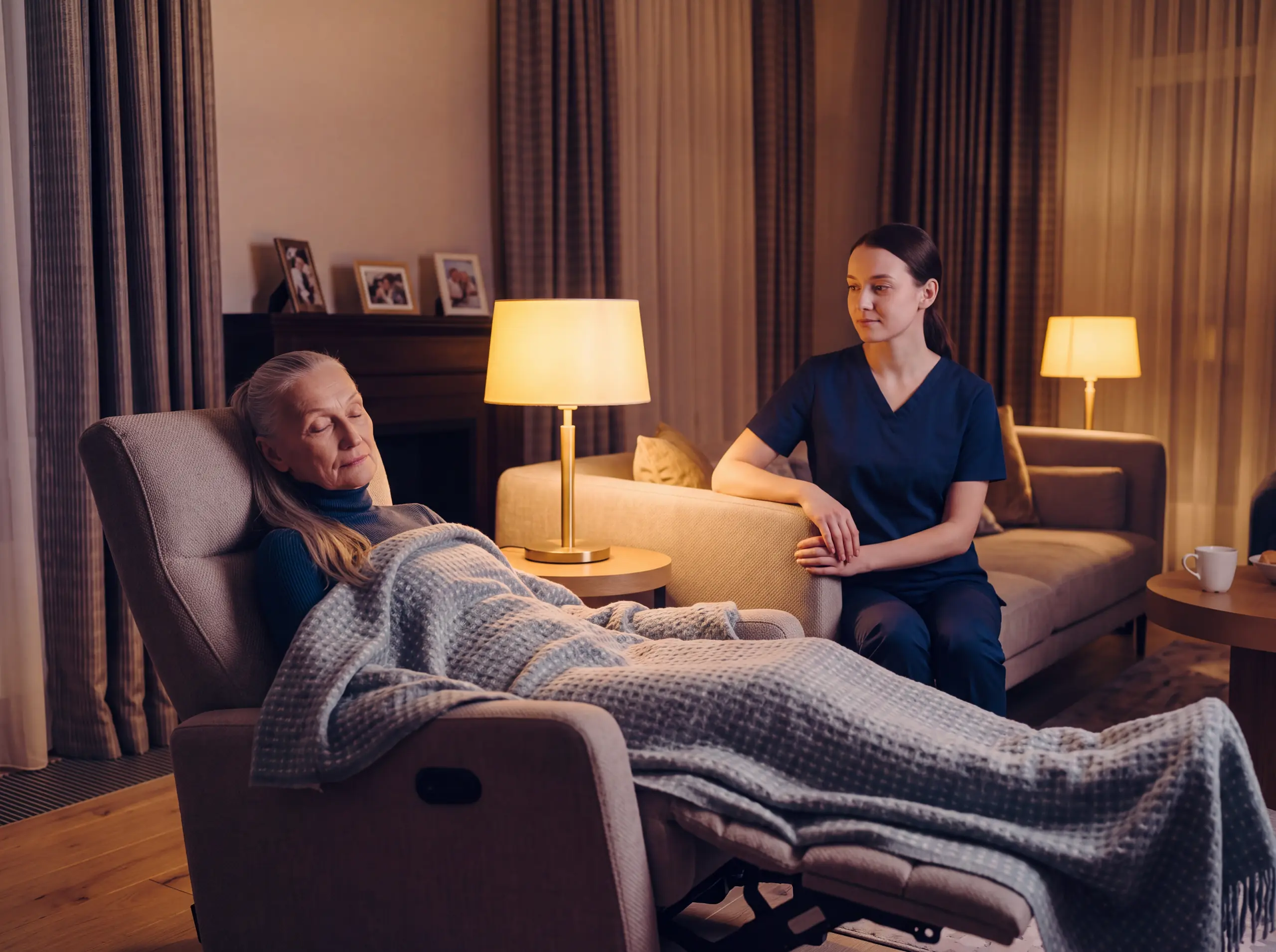 Caregiver sits beside a senior woman resting under a blanket in a recliner in a cozy, lamp-lit living room.