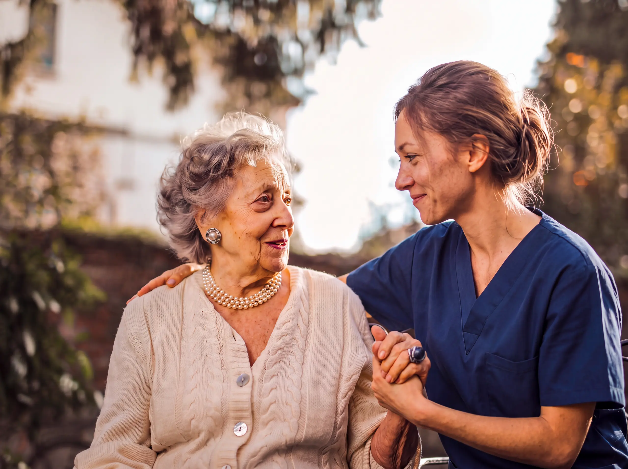 Smiling caregiver in navy scrubs holds hands with a senior woman outdoors, offering companionship.
