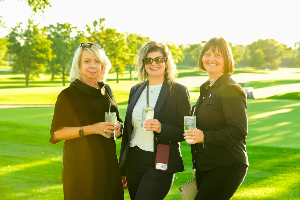 Three European Service team members smiling with drinks on a sunny course at a community golf outing.