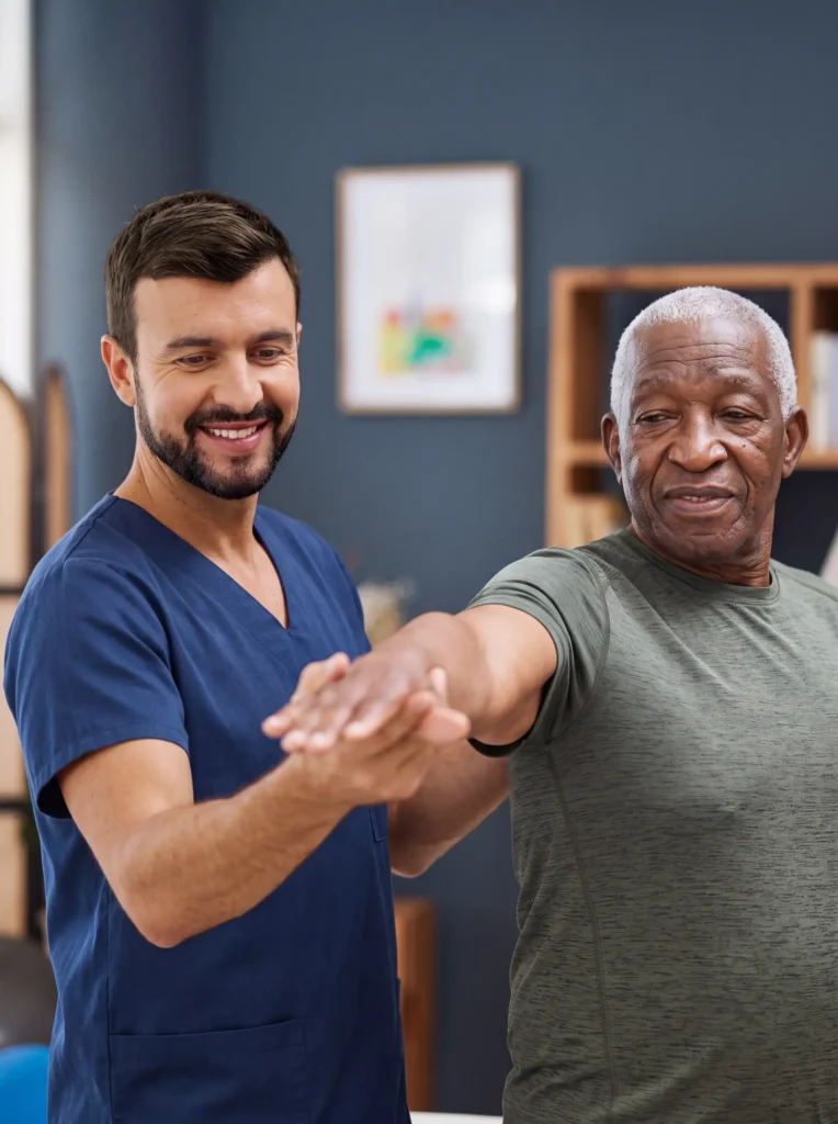 Male caregiver in navy scrubs supporting a smiling senior man's outstretched arm during a gentle stretching exercise at home.