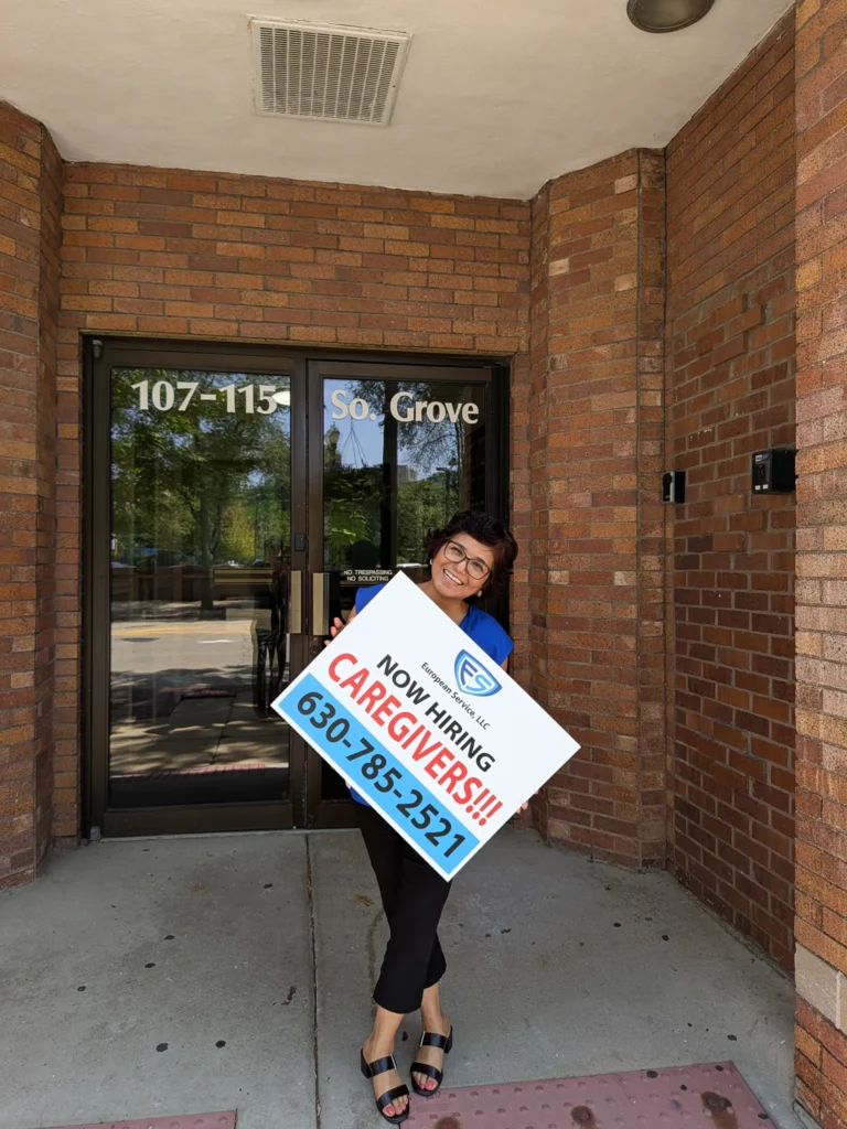 Team member holding a “Now Hiring Caregivers” sign at the 107–115 S Grove Avenue entrance of the European Service office in Elgin, Illinois.